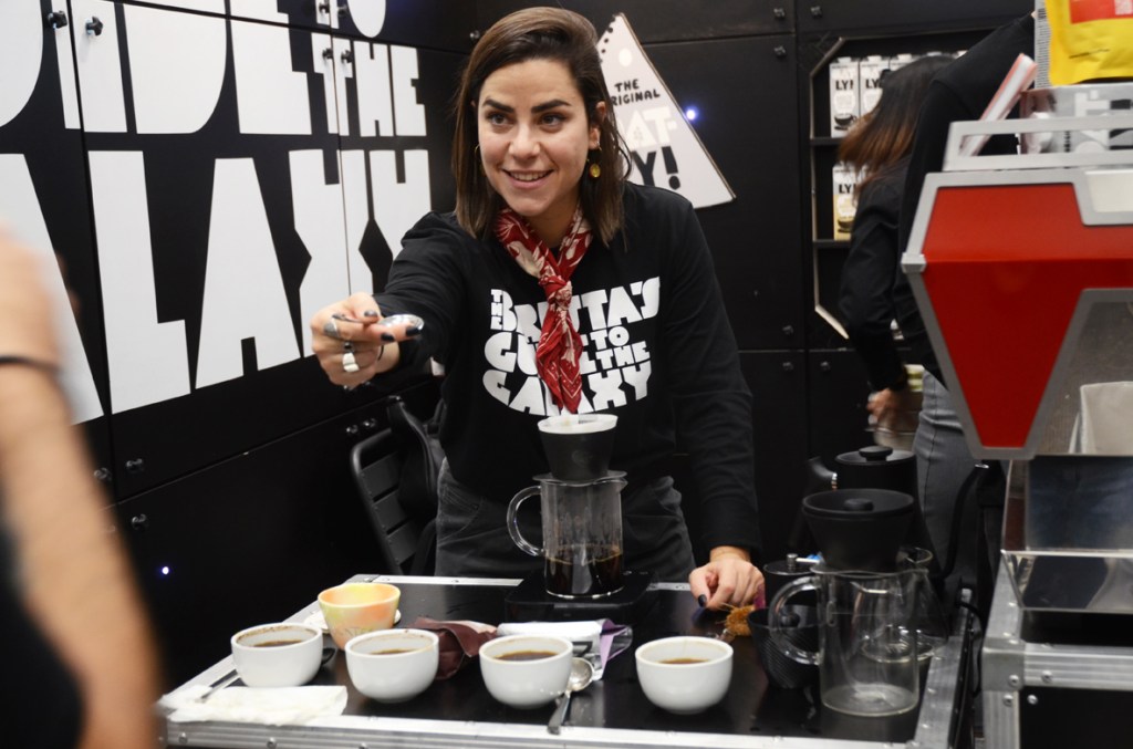 Young woman offering a coffee cupping spoon to someone over a table filled with brewing coffee