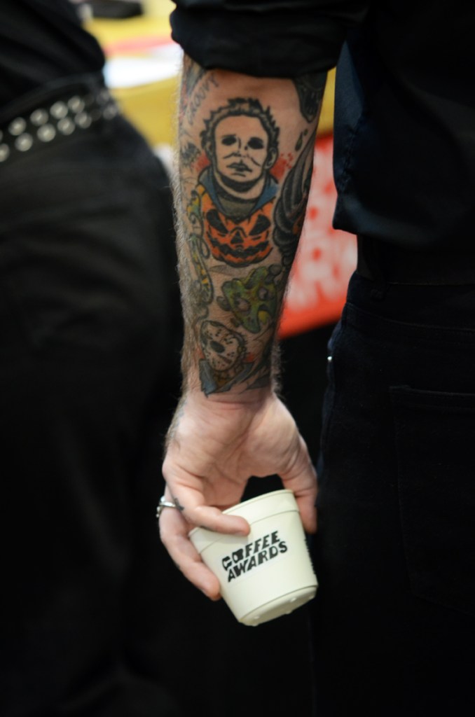 A Michael-Myers-and-Halloween-pumpkin-tattooed arm with hand clutching Barcelona Coffee Awards coffee cup