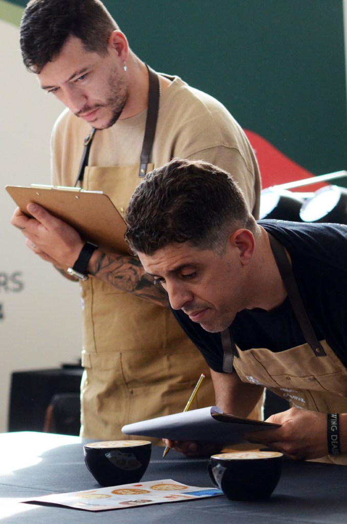 Latte art judge peering over two recently poured lattes with a clipboard and pencil in hand